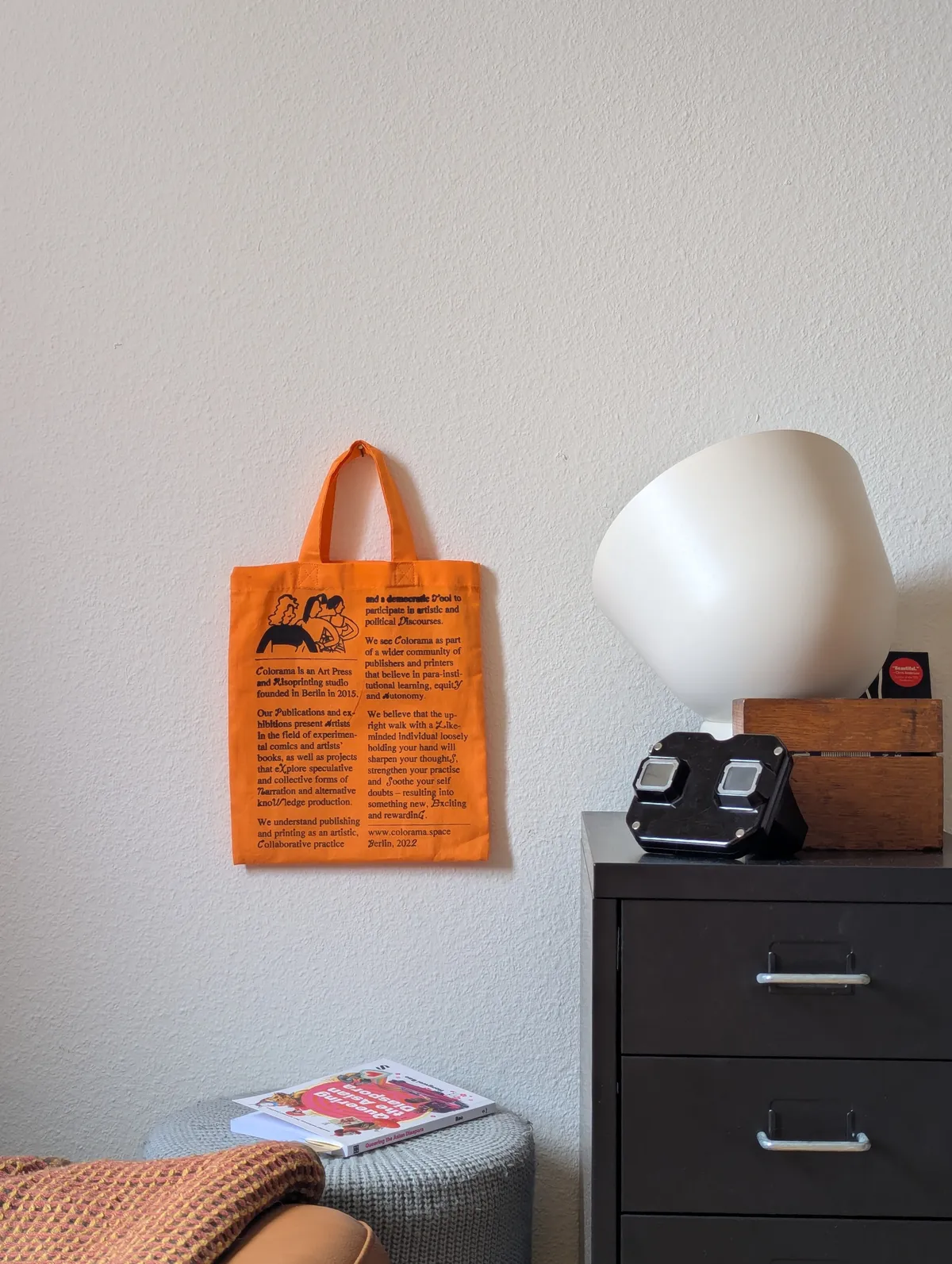 A bright orange book bag hangs from the wall next to a black metal drawer unit with a black View-Master on top.