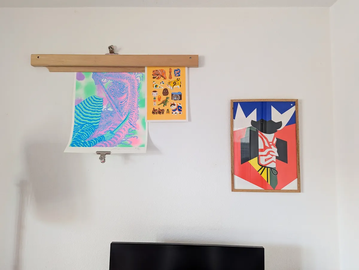 A white wall with a gravity bar holding two art prints. A framed graphic Illustration of a samurai hangs next to it.