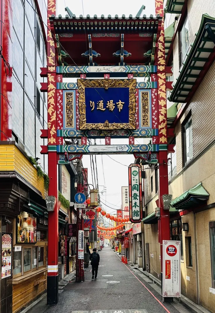 A colorful archway marks the entrance to a bustling street in a traditional market area, lined with shops and restaurants. Lanterns hang overhead, and a person walks down the narrow path surrounded by vibrant signage.