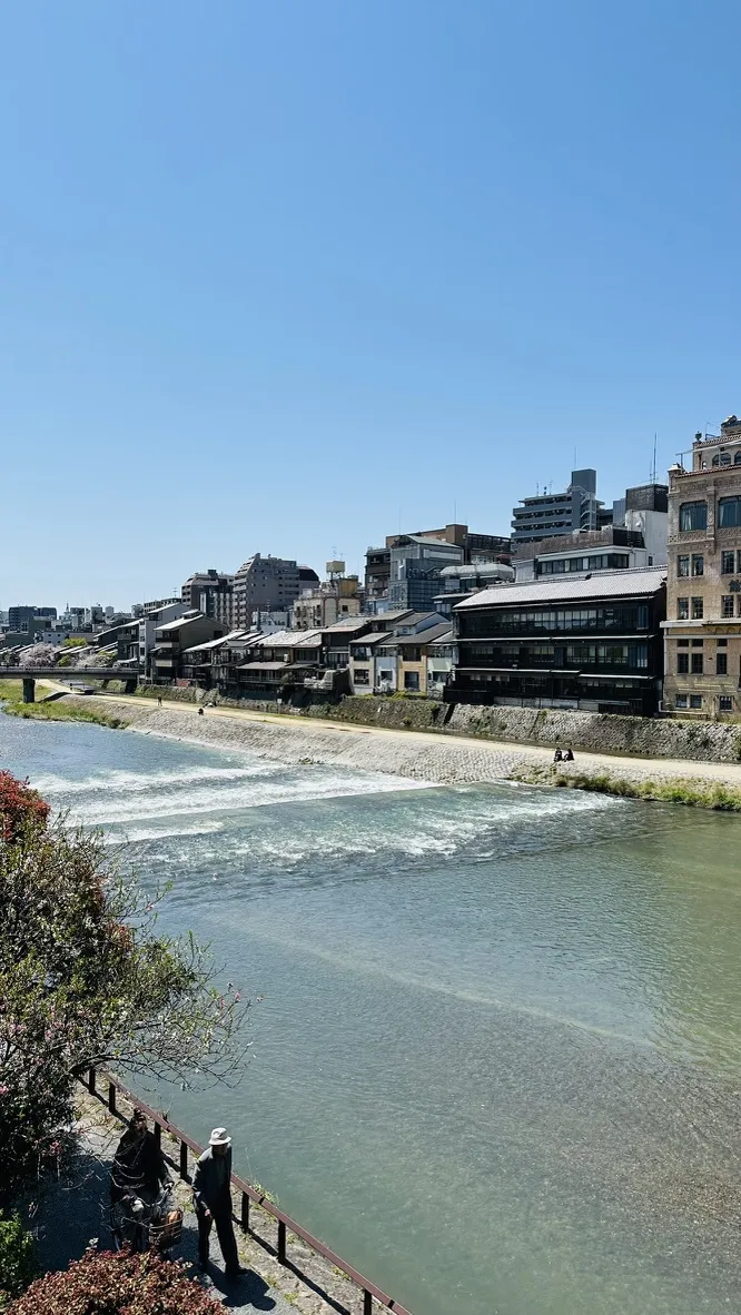 A serene river flows alongside a cityscape under a clear blue sky. Buildings line the riverbank, with a mixture of modern architecture and traditional structures, while people can be seen enjoying the view from the edge.