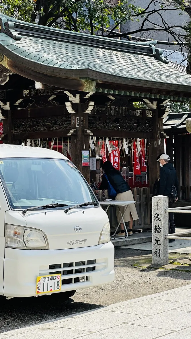 A small white van is parked in the foreground while a person in a skirt is engaged in an activity at a Shinto shrine in the background, where red and white banners hang. A stone marker is visible to the right, indicating the shrine's name.