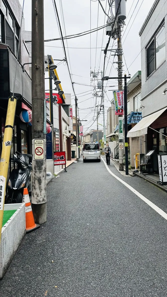 A narrow street in an urban area featuring power lines overhead, a parked motorcycle on the left, and pedestrians walking. Various businesses are visible along the sides of the street, with signs in Japanese.