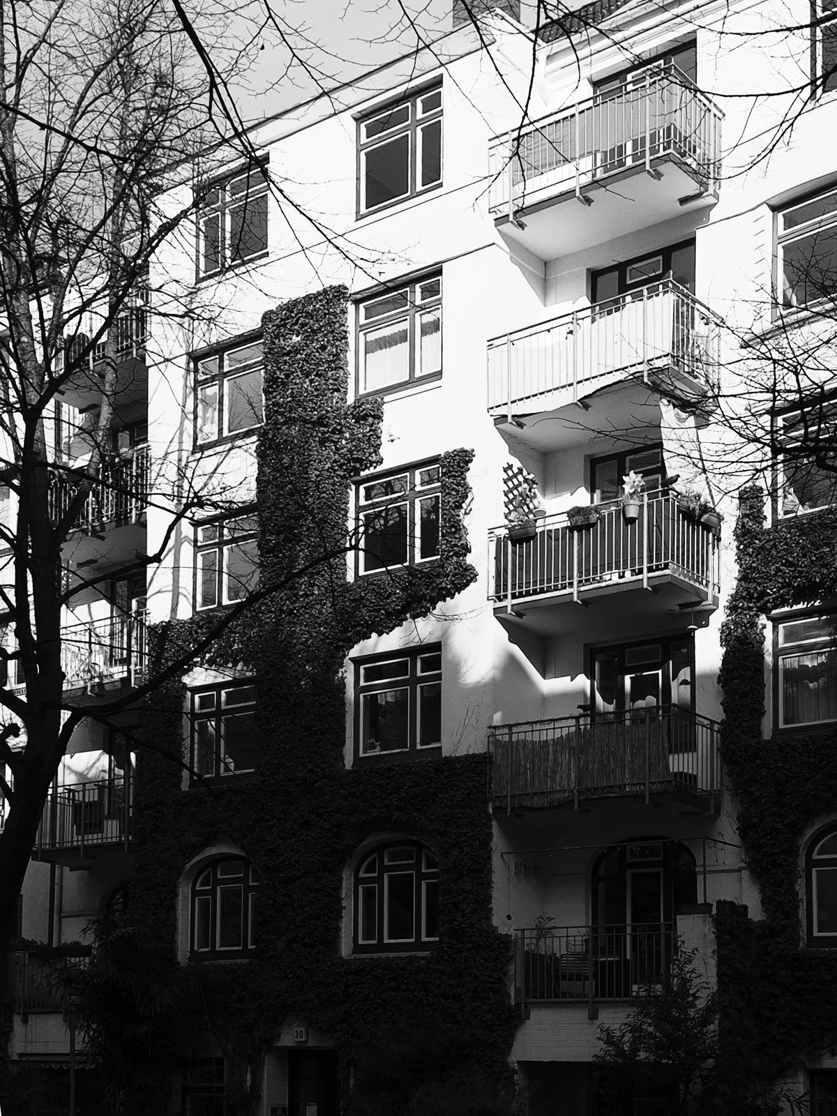 A multi-story residential building partially covered in ivy, featuring several balconies and large windows. The image is in black and white, with stark contrasts created by sunlight and shadows.