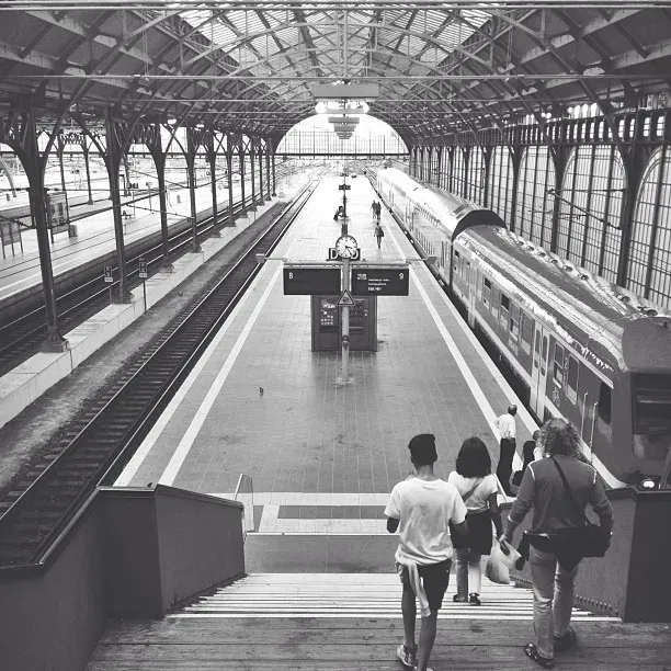 A black-and-white photo of a railway station featuring empty platforms and a large clock. People walk down the stairs toward the platform, where a train is parked and the station has high ceilings supported by iron beams.