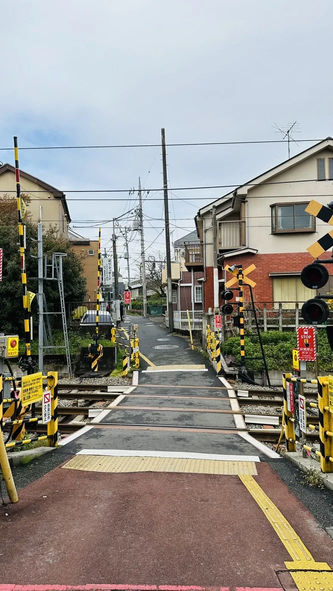 A narrow pathway leads to a railway crossing, with yellow and black warning signs and lights on either side. Residential buildings are visible in the background, along with overhead power lines and a cloudy sky.