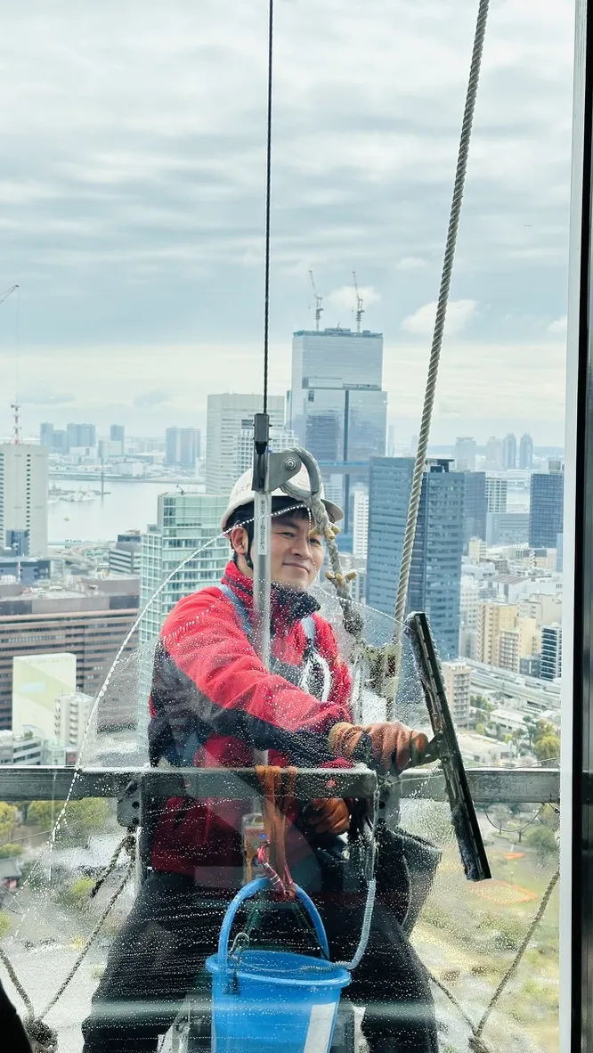 A window cleaner, wearing a helmet and harness, is suspended on a platform while cleaning large glass windows. The city skyline, including tall buildings and a body of water, is visible in the background under a cloudy sky.