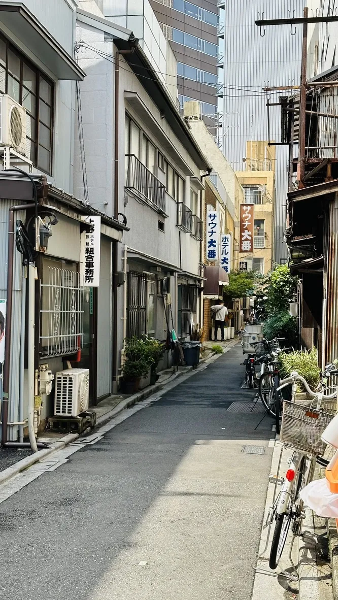 A narrow urban alleyway lined with traditional buildings and storefronts, featuring hanging banners and a bicycle parked on the right. The scene captures a blend of modern and historic architecture, with high-rise buildings visible in the background.