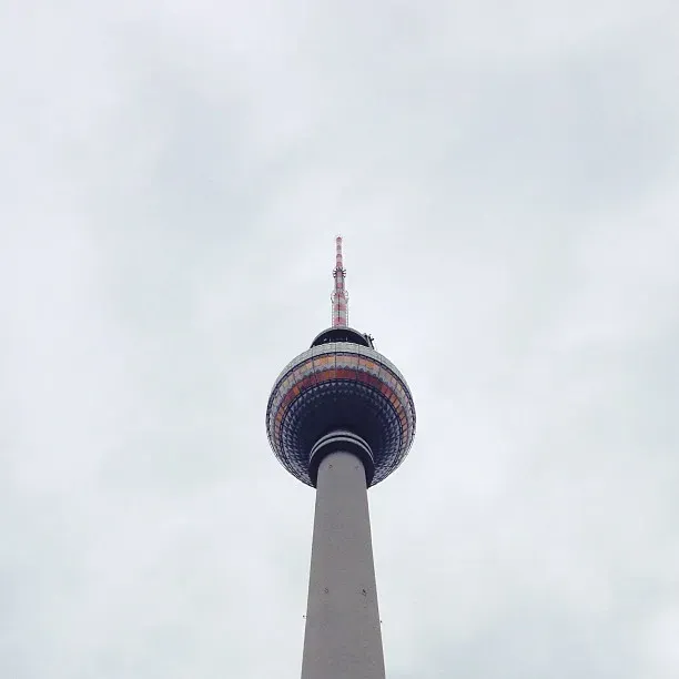 A view looking up at a tall television tower with a round observation deck and antenna on top, set against a cloudy sky. The tower features colorful rings around its midsection.