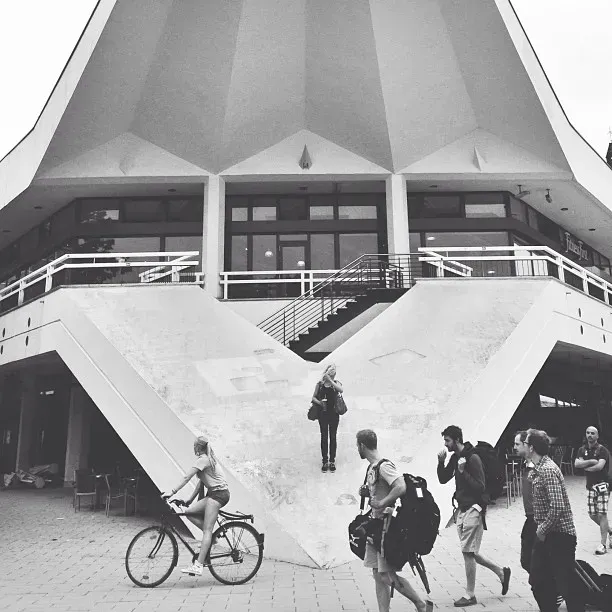 A modern building with a distinctive angular design features a wide staircase at its entrance. In the foreground, a person rides a bicycle while others walk nearby, creating a lively atmosphere.