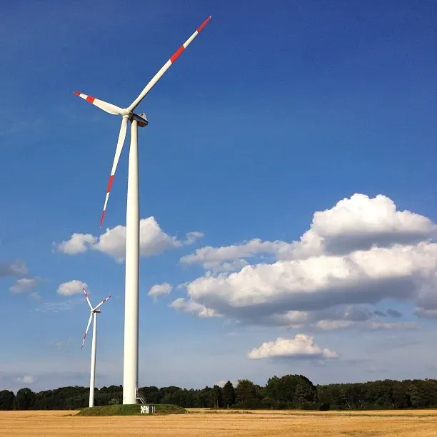 Two tall wind turbines stand against a blue sky adorned with fluffy white clouds, set in a field with golden crops. The turbines have red and white striped blades and are surrounded by trees in the background.