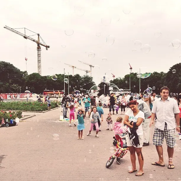 A bustling park scene filled with families and children enjoying a day out. People are seen playing with bubbles, while children ride bicycles and others are gathered in various activities, with construction cranes visible in the background.