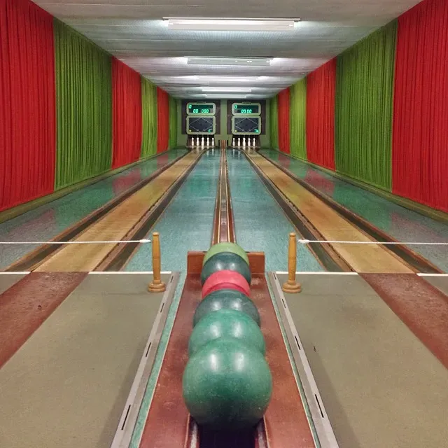 A view down a bowling lane featuring three bowling balls in the foreground, with striped red and green drapes lining the walls. The pins are set at the end of the lane, and scoring monitors are visible above the pins.