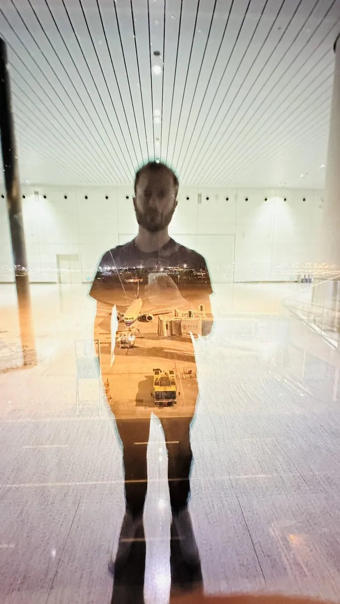 A person stands in front of a glass window, creating a reflection that blends their silhouette with a view of an airport tarmac and grounded airplanes at night. The surroundings appear minimalistic with bright overhead lights.