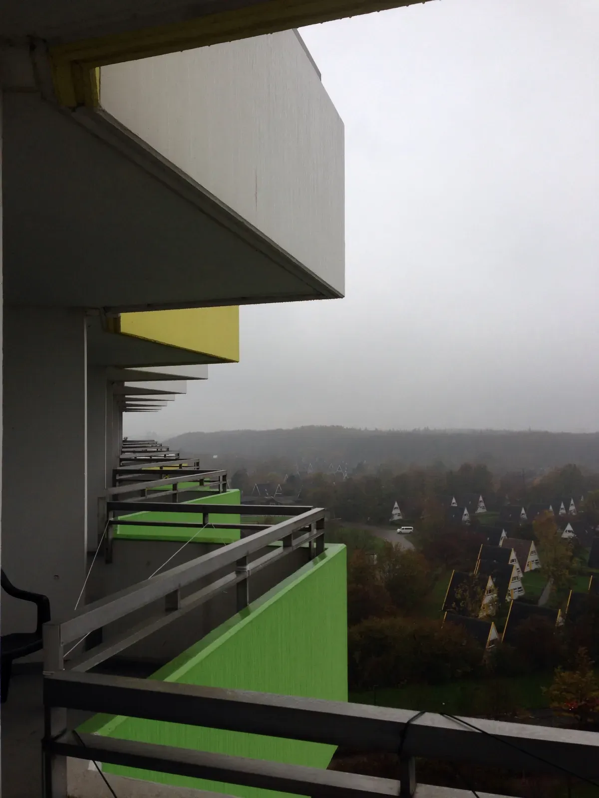 Balconies with vibrant yellow and green accents protrude from a multistory building, overlooking a misty landscape filled with trees and a series of triangular-roofed houses in the distance. The weather appears cloudy and gray, creating a subdued atmosphere.
