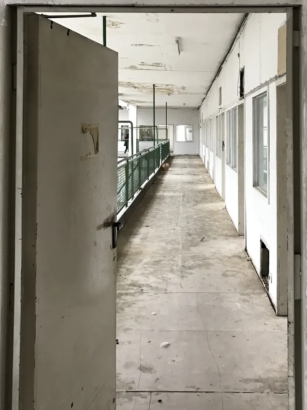 A door is ajar, revealing a long, narrow hallway with faded white walls and a worn floor. Windows line the sides, and a green railing runs alongside the corridor, which appears empty and unkempt.