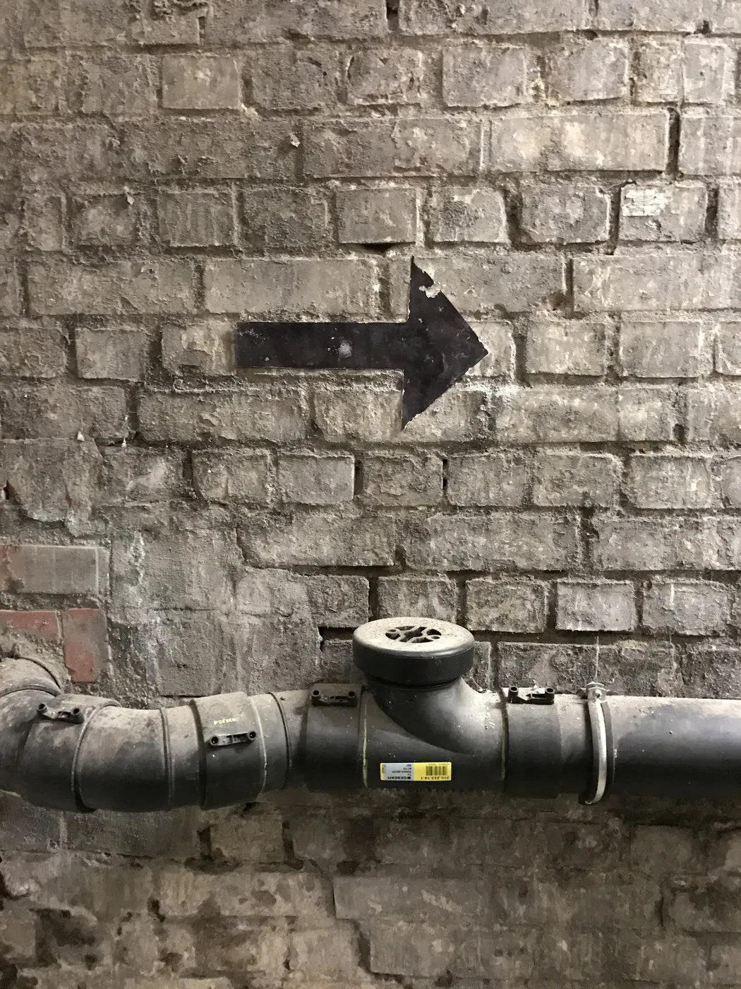 A dirt-stained brick wall features a large, black arrow pointing to the right. A dark, curved pipe runs horizontally along the bottom of the image.