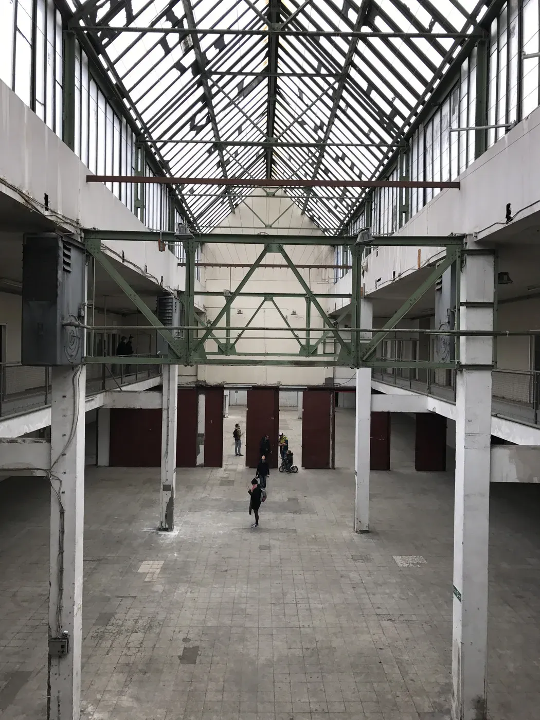 An empty industrial space with high ceilings and large windows, showcasing a grid of steel beams above. Sparse concrete flooring is visible, and a few people are walking in the center, with red doors at the far end of the hall.