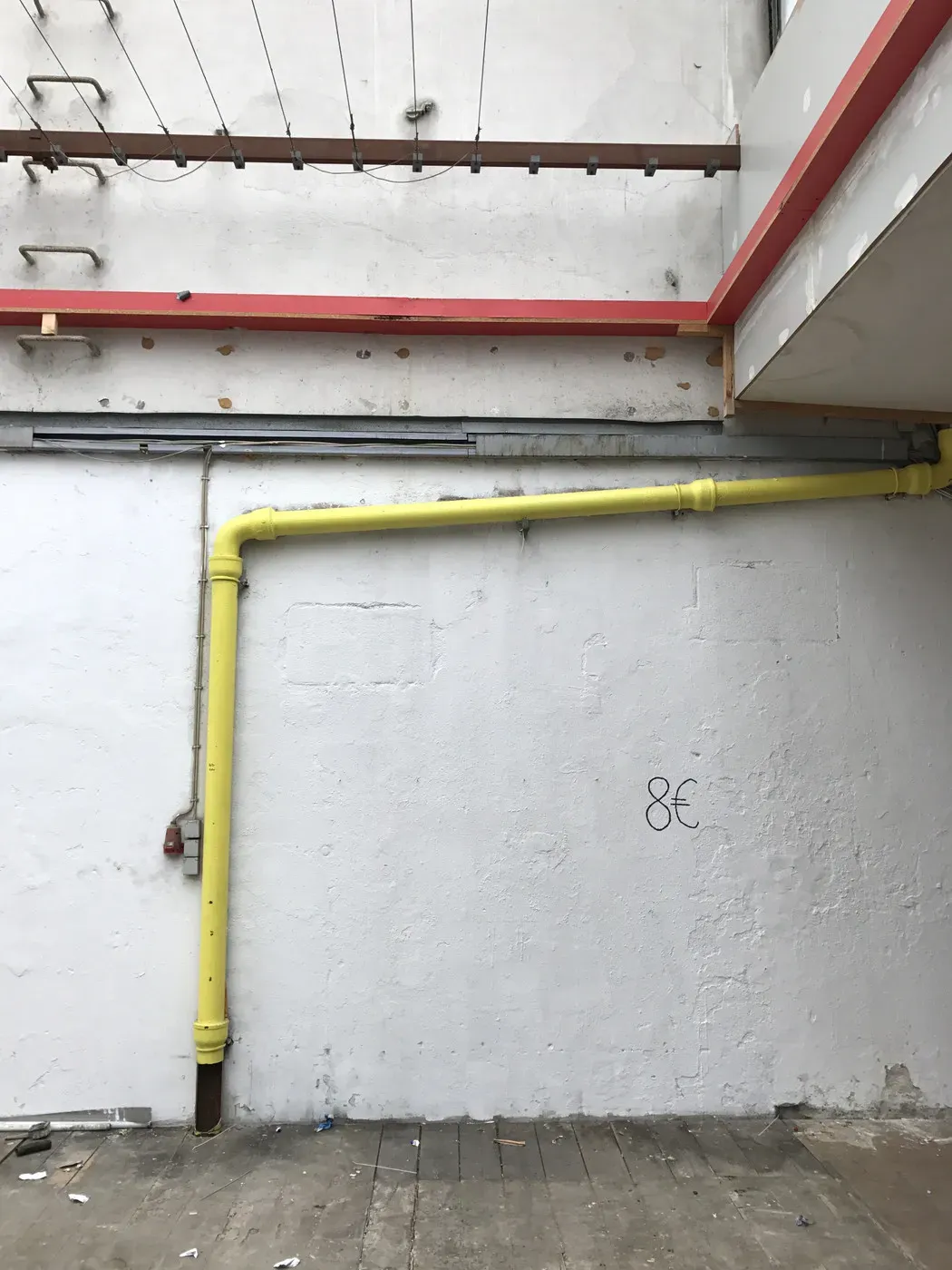A wall with a rough white surface features a prominent yellow pipe running diagonally downward. Above, there is a section of exposed wooden beams and a red strip along the ceiling. The floor is made of wooden planks, and a faint price tag reading '8€' is visible on the wall.