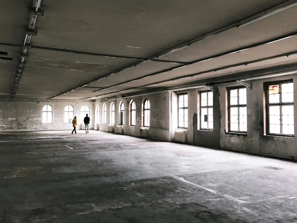 Two figures walk across a spacious, empty room with large arched windows. The walls are bare and the floor is grey concrete, creating an industrial ambiance.