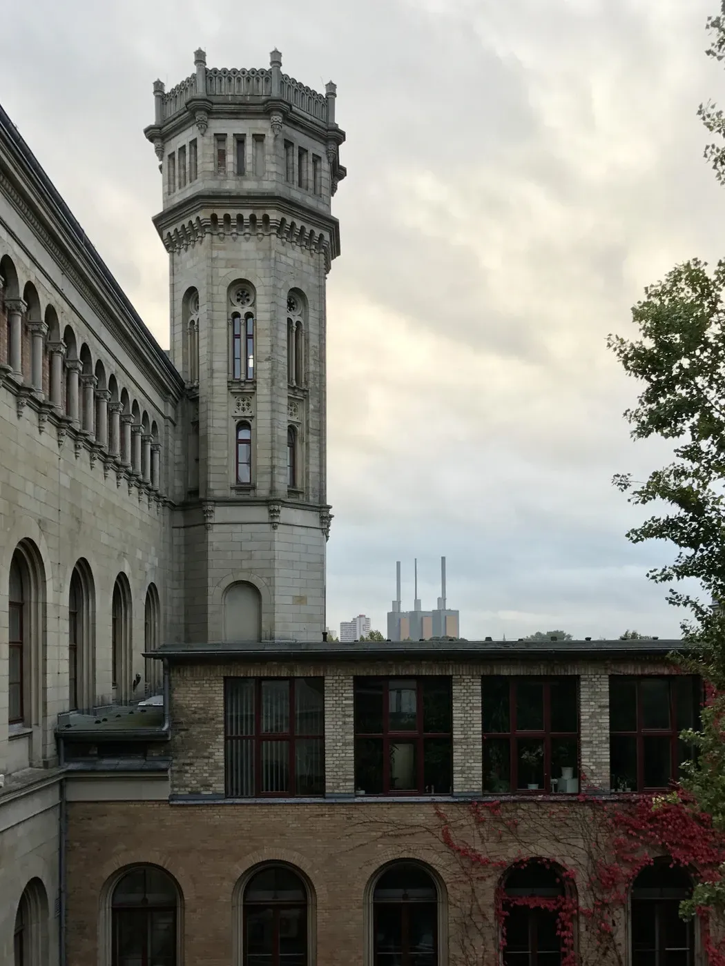 A historic building with a tall, ornate tower is contrasted by a modern skyline in the background, featuring industrial structures. The scene is set under a cloudy sky, with greenery partially framing the foreground.