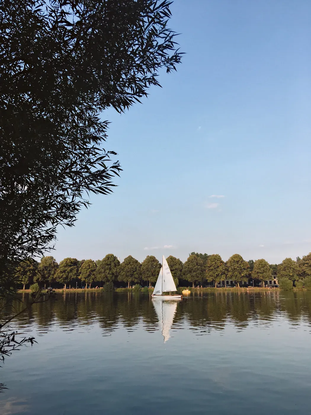 A tranquil lake scene featuring a small sailboat gliding on the water, surrounded by lush greenery and trees reflecting in the calm surface. The sky above is clear with soft clouds.