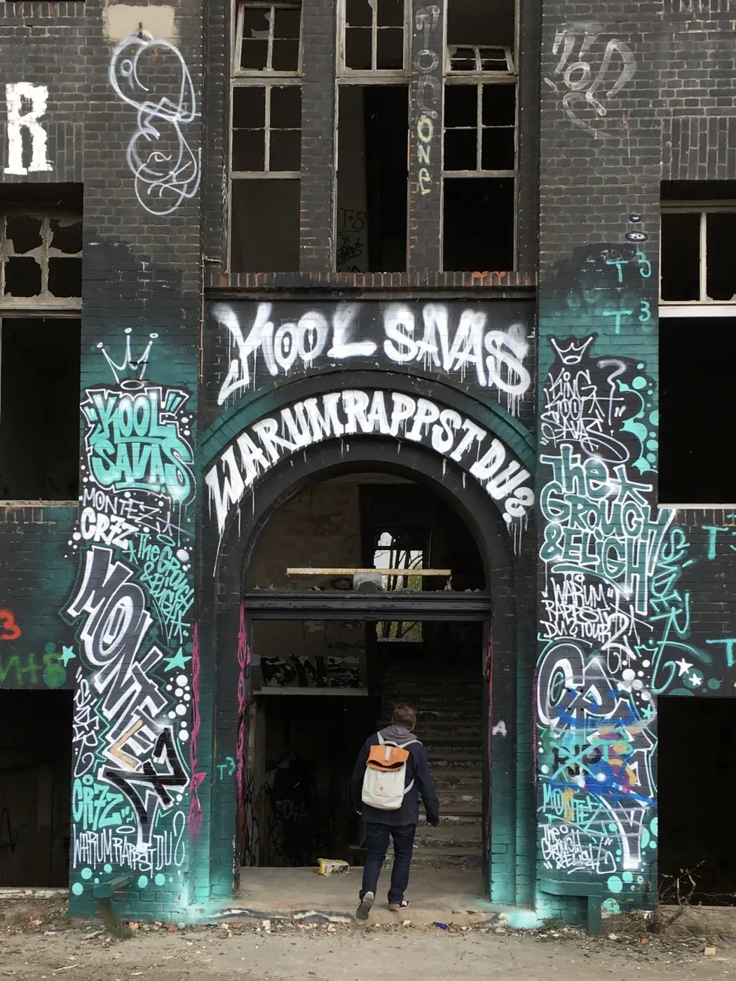 A person with a backpack walks through an arched entrance covered in colorful graffiti, featuring words like 'Kool Savas' and 'Marvins Rapstadium.' The surrounding brick walls are adorned with various tags and designs.