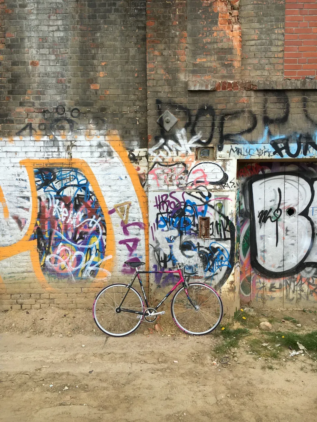 A single bicycle with pink accents stands against a vibrant graffiti-covered wall. The wall features various colors and designs, including tags and stencils, alongside exposed brick and a partially visible door.