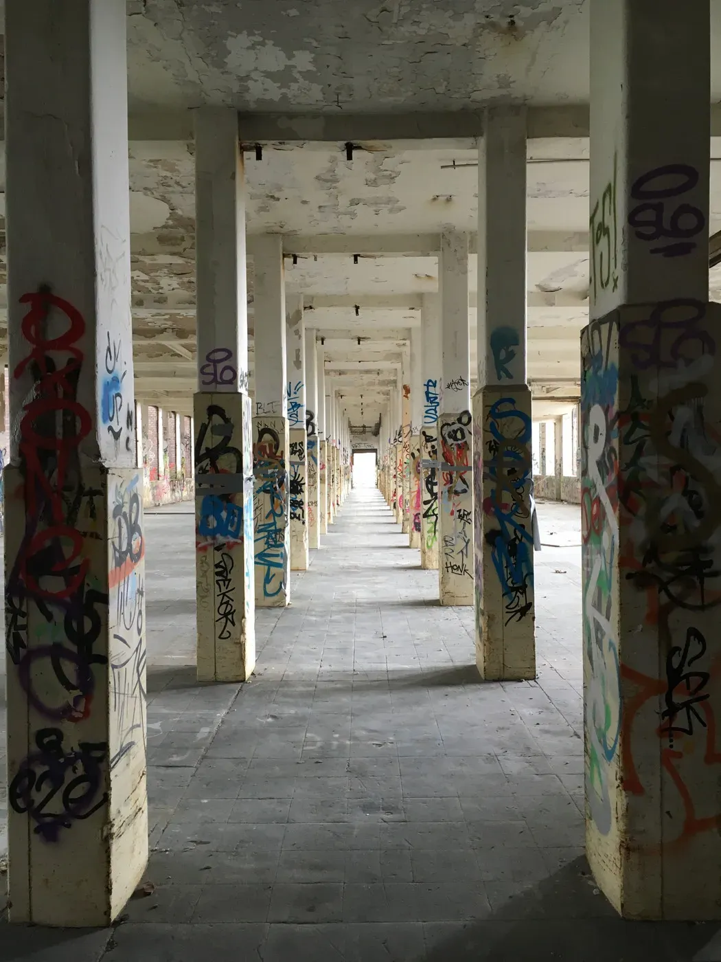 A long corridor lined with tall, white pillars covered in colorful graffiti. The floor is tiled and the walls show signs of decay, with peeling paint and an open space at the far end leading to light.