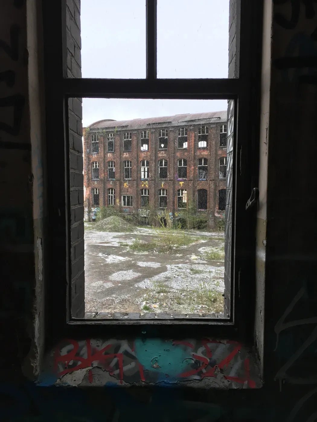 A rusted window frame reveals a view of a dilapidated brick building with broken windows, surrounded by overgrown vegetation and debris in the foreground. Graffiti is visible on the walls around the window.