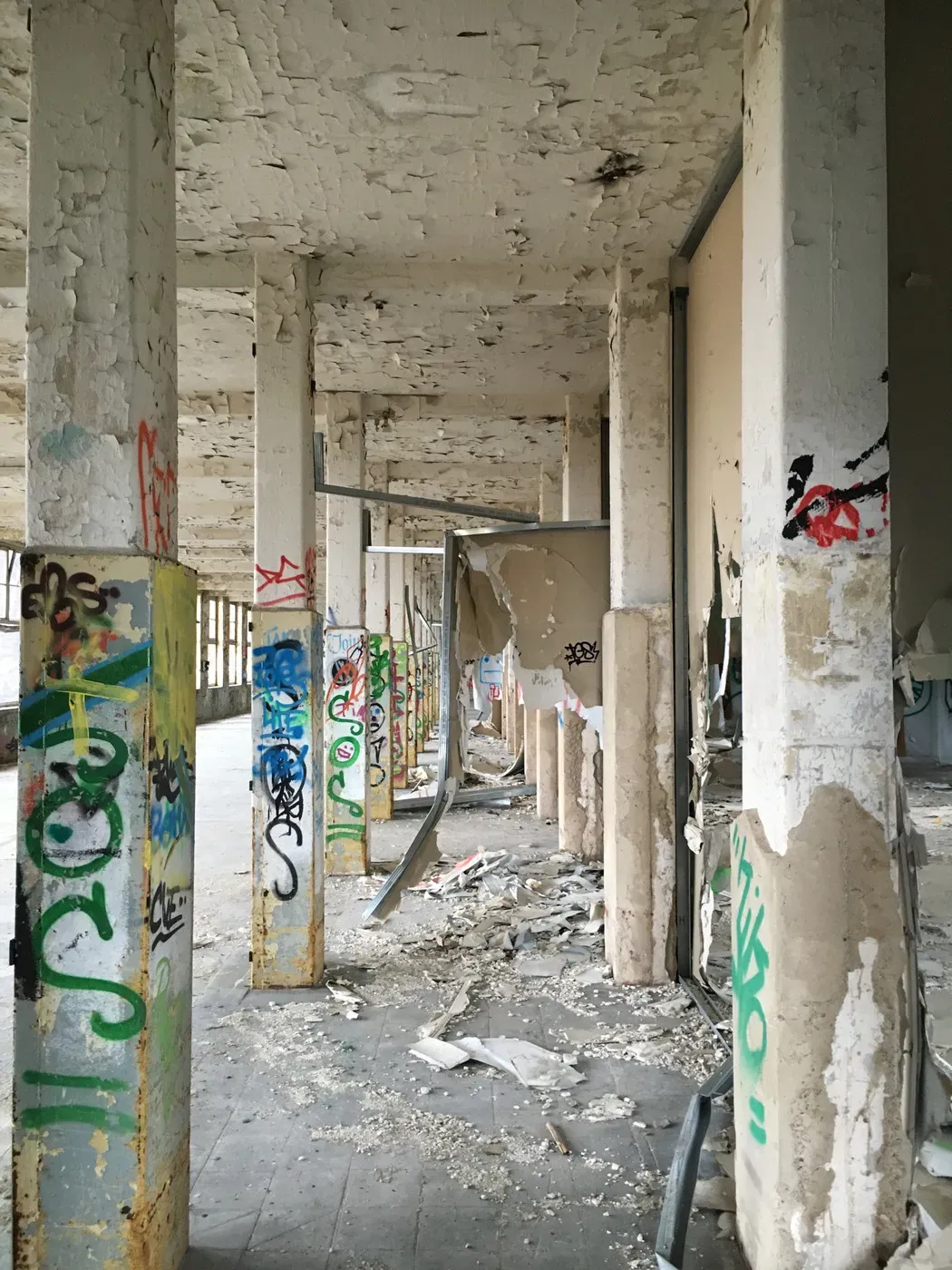 A corridor with deteriorating columns shows peeling paint and colorful graffiti along the walls. Debris and litter cover the floor, contributing to the abandoned and neglected atmosphere.
