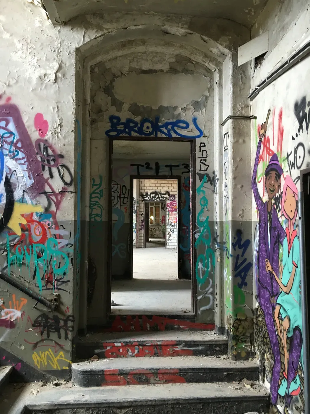 A view through an abandoned hallway with peeling walls covered in colorful graffiti. The corridor leads deeper into the building, showing more graffiti on the walls and a sense of decay.