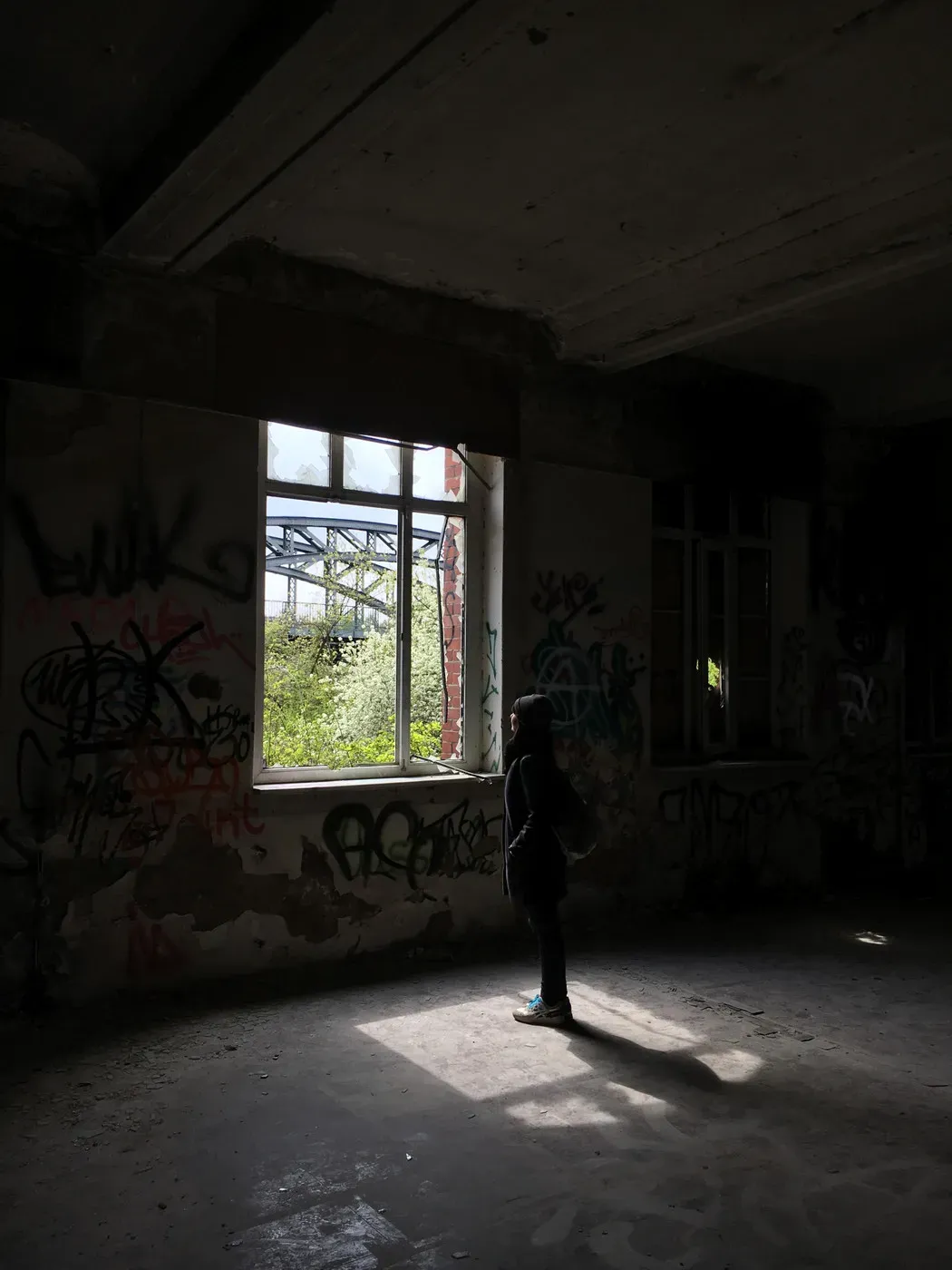 A silhouette of a person stands in a dimly lit room, facing a large, partially open window. The walls are covered in various graffiti, and greenery is visible outside the window.