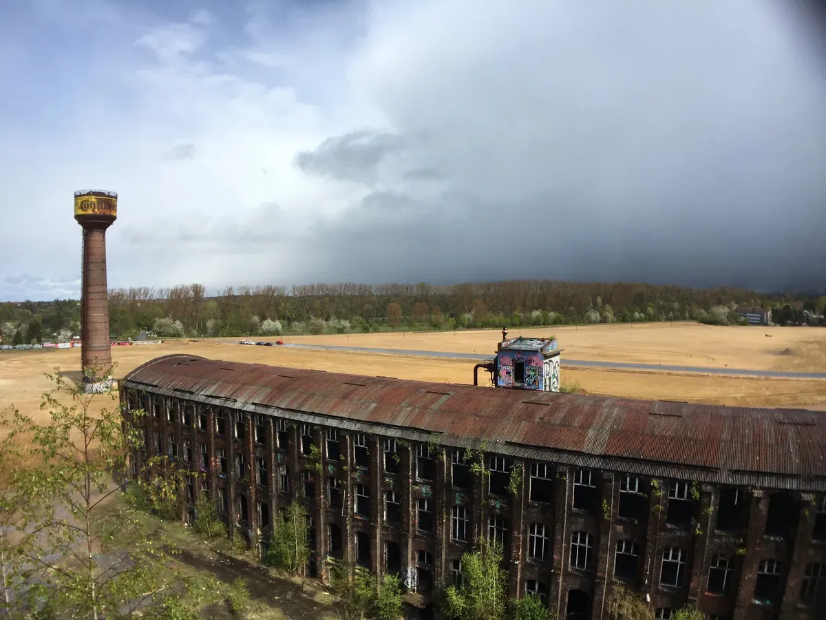 A tall, weathered water tower stands next to an abandoned, dilapidated industrial building with a rusted roof. The landscape features a field with sparse greenery and dark storm clouds gathering in the distance.