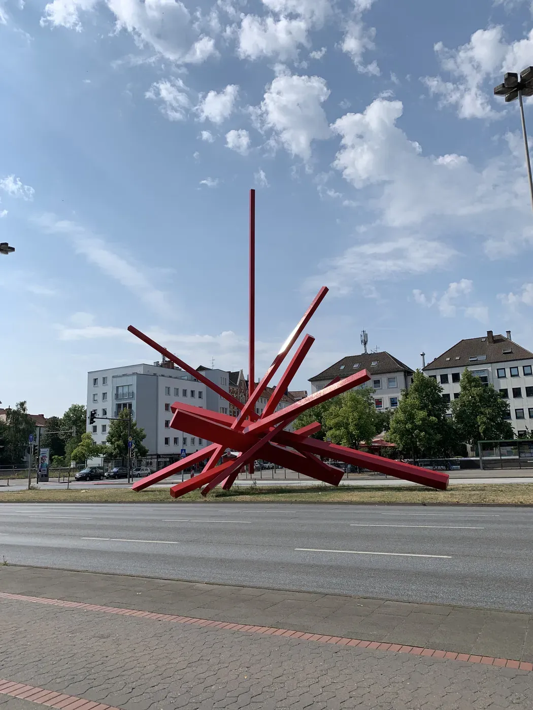 A large, abstract red sculpture made of intersecting beams is positioned in a grassy area near a road. The background features buildings and a blue sky with scattered clouds.