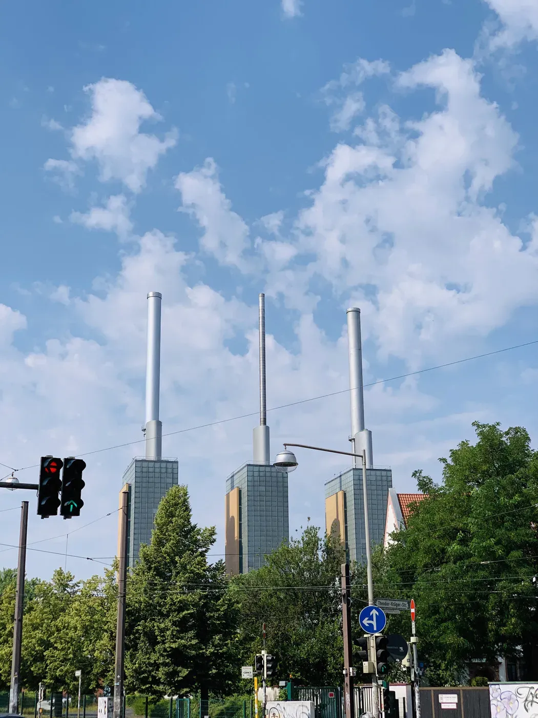 Three tall industrial smokestacks rise from a modern building with glass facades against a backdrop of a blue sky with scattered clouds. Green trees and traffic lights are visible in the foreground.