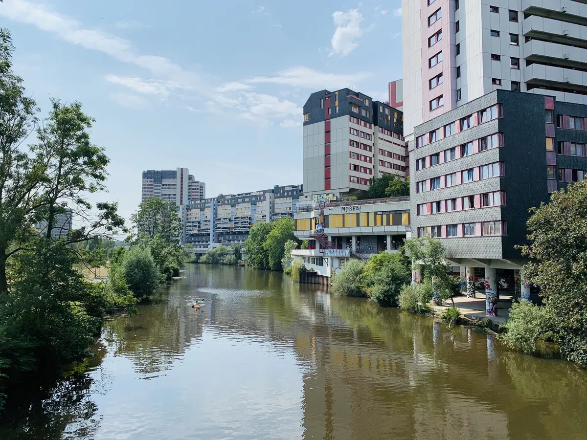 A calm river runs through an urban area, flanked by modern buildings and greenery. Two people in a kayak are paddling on the water, under a clear blue sky with scattered clouds.
