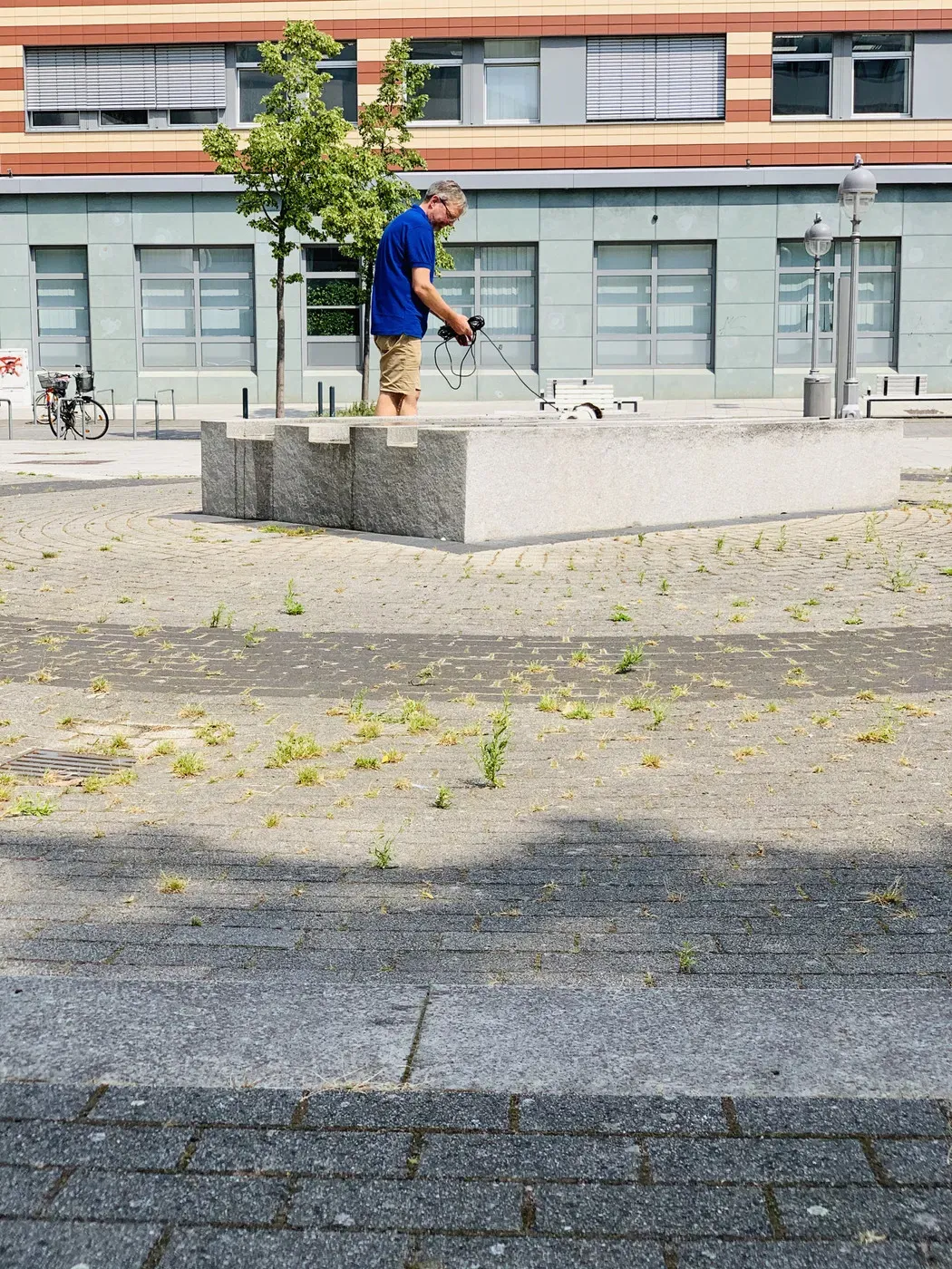 A man in a blue shirt stands on a stone slab in a mostly empty plaza, holding a device and looking down. Surrounding him are patches of grass and small plants growing through the stone pavement, with buildings and bicycles visible in the background.