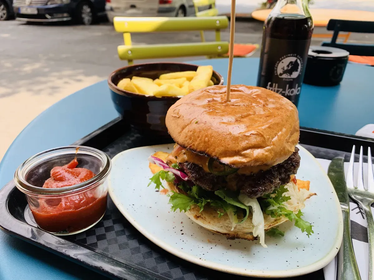 A gourmet burger on a plate, topped with fresh lettuce and served with a side of crispy fries in a bowl. A small jar of sauce is placed alongside, and a bottle of fizzy drink is visible in the background on a blue table.
