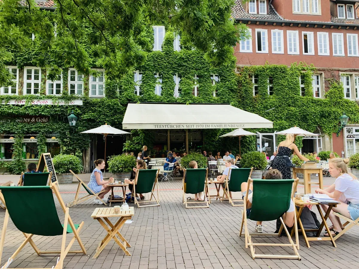 A lively outdoor seating area with green lounge chairs and tables under parasols, surrounded by lush ivy-covered buildings. People are enjoying drinks and engaging in conversation in a relaxed atmosphere.