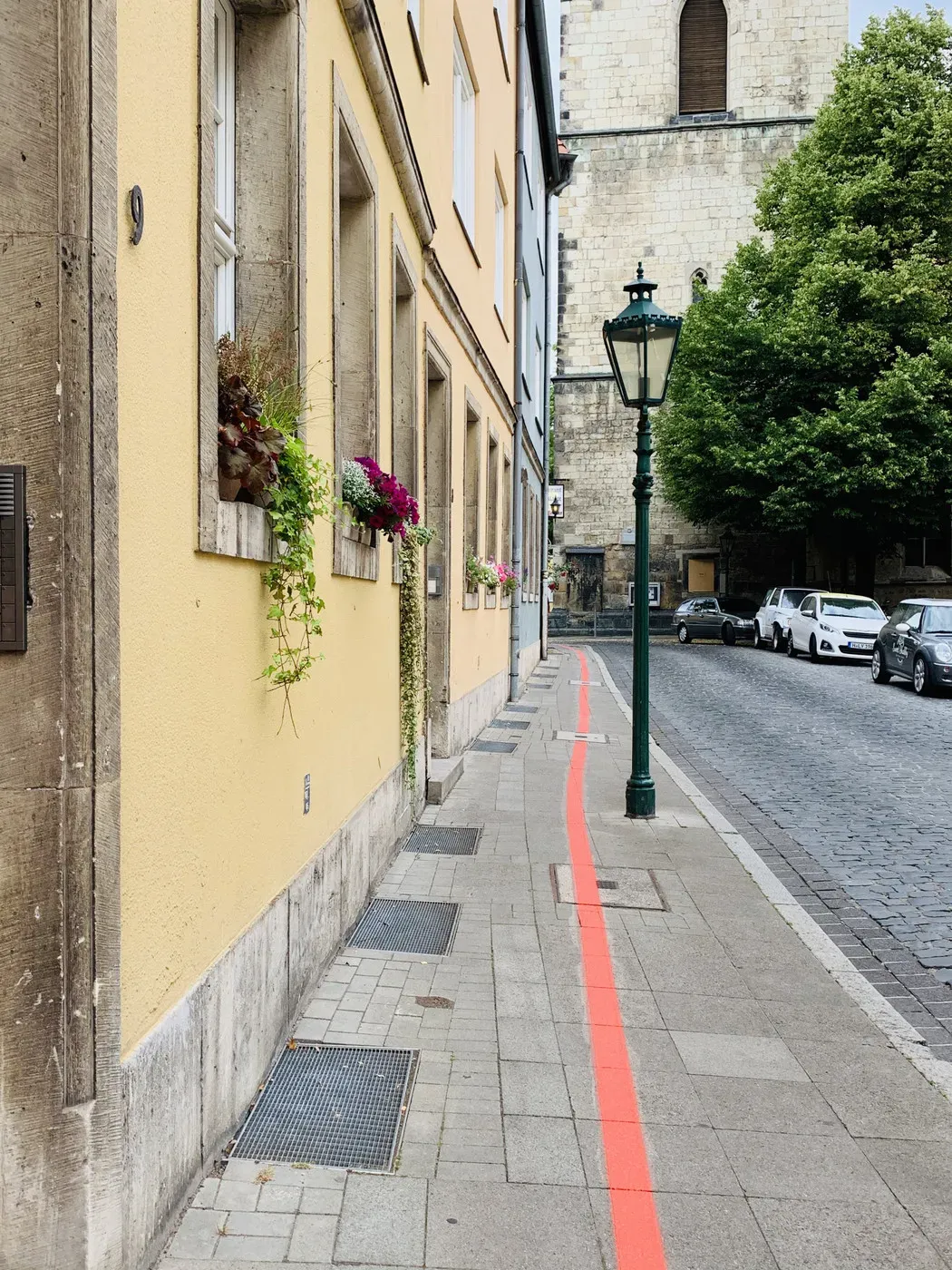 A narrow street lined with a yellow building featuring flower boxes beneath its windows. A red-painted line runs along the pavement, leading towards a green street lamp, with a stone church visible in the background.