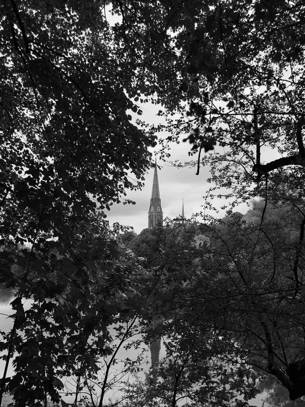 A view through trees frames a tall church spire rising above the landscape, with water reflecting the surrounding scenery in black and white tones.