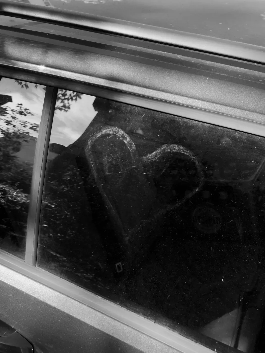 A heart shape is drawn in the dust on a car window, with reflections of trees and surroundings visible in the glass. The image is in black and white.