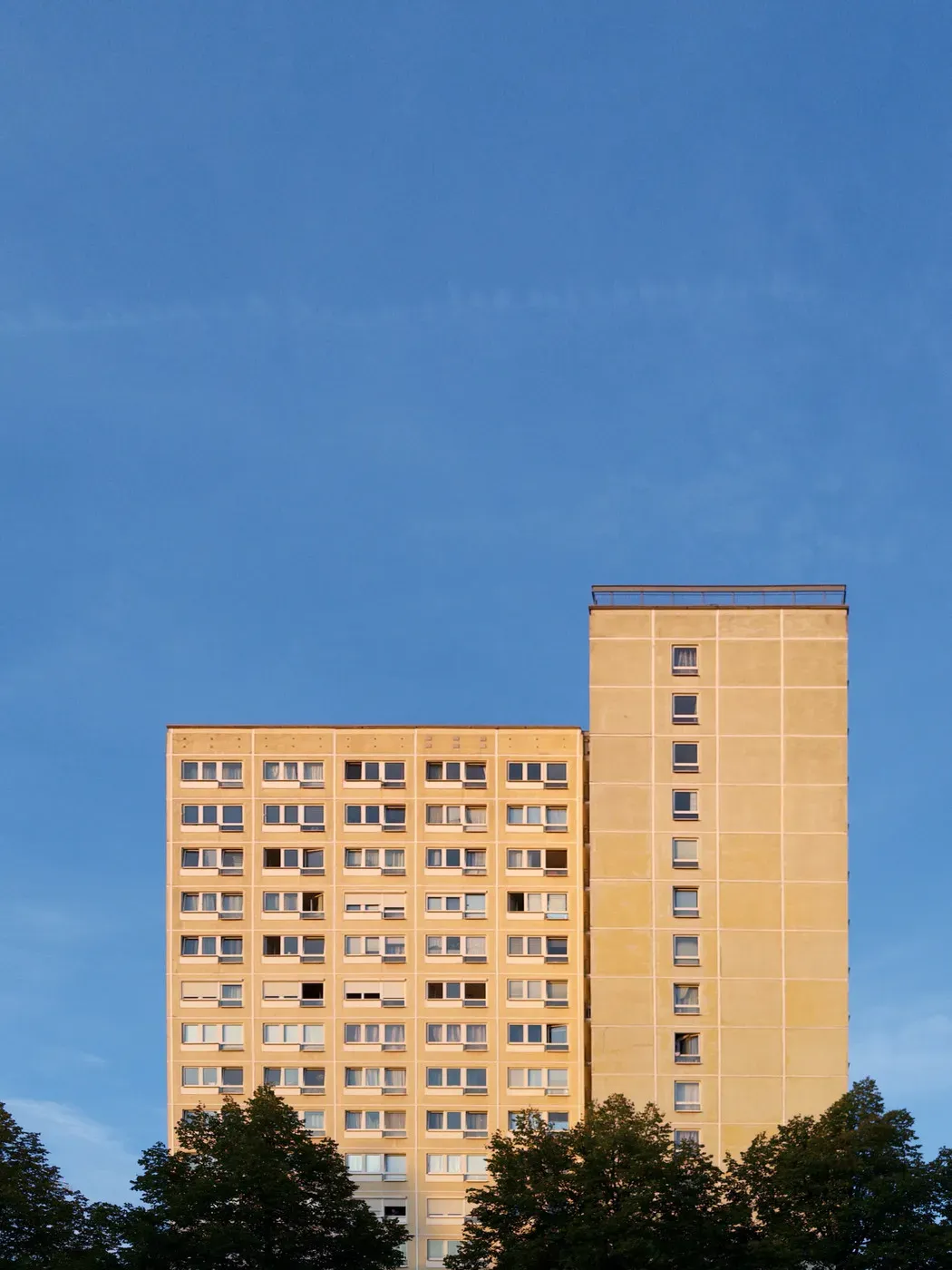 A tall, yellow apartment building stands against a clear blue sky, featuring multiple windows and a distinctive vertical structure. The lower section has a wider base, while the upper portion is narrower.