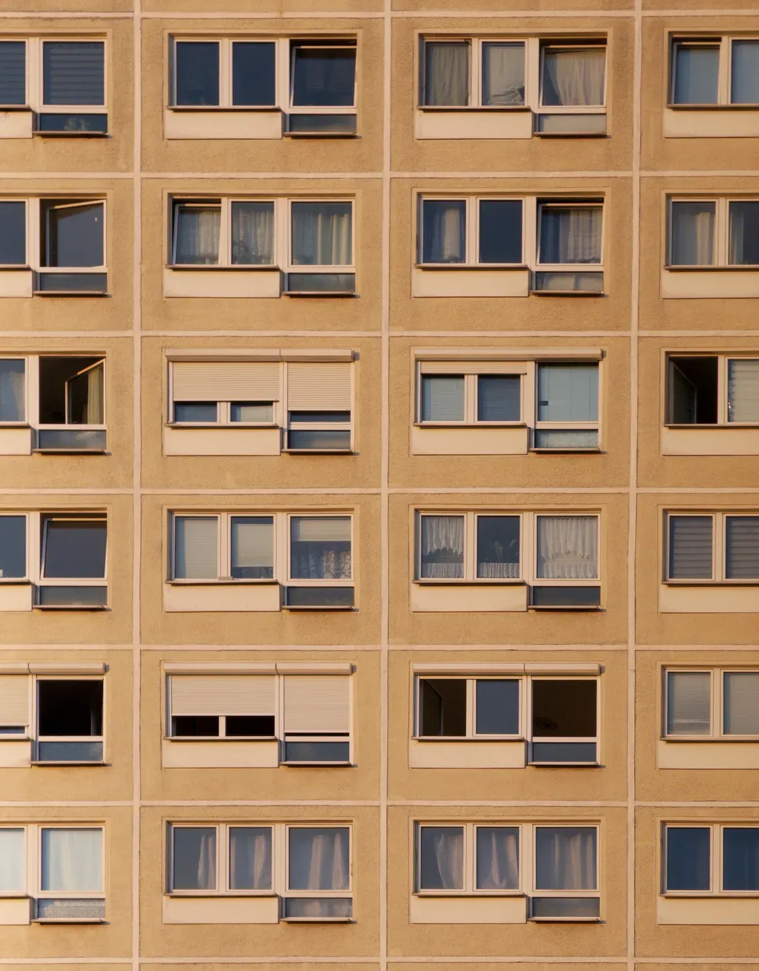 A close-up view of a building's façade featuring a grid of windows. Many windows are adorned with sheer curtains, while some are partially open or closed, revealing a uniform beige exterior wall.