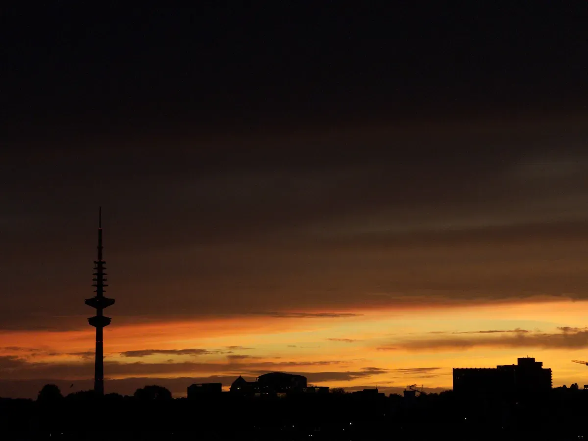 A skyline is silhouetted against a dramatic sunset, featuring a tall communication tower and various structures. The sky displays rich hues of orange and purple as the daylight fades.