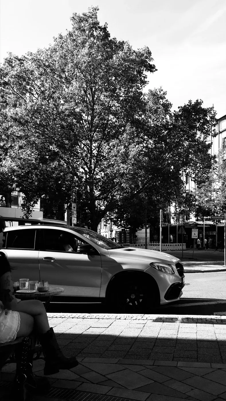 A silver car is parked on a street, partially obscured by a large, leafy tree. In the foreground, a person sits at a café table, wearing white shorts and black boots.