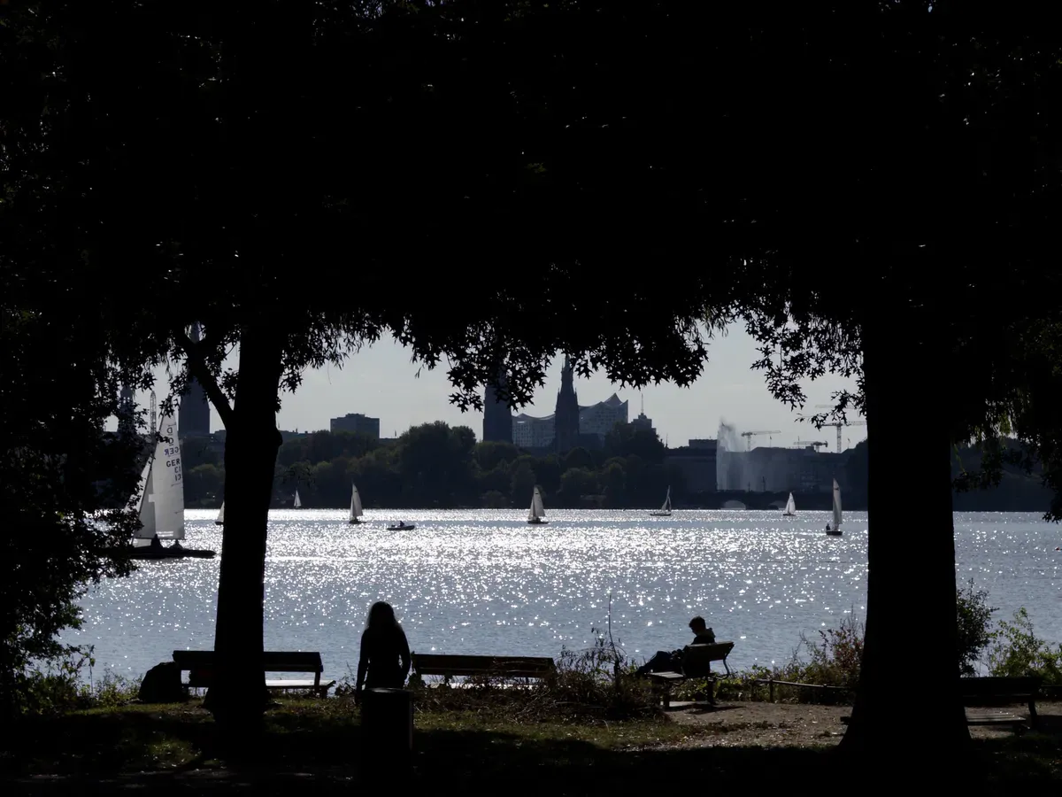 A serene lake sparkles in the sunlight, with small sailboats gliding across its surface. Silhouetted trees frame the scene, and distant city buildings are visible against a bright sky.