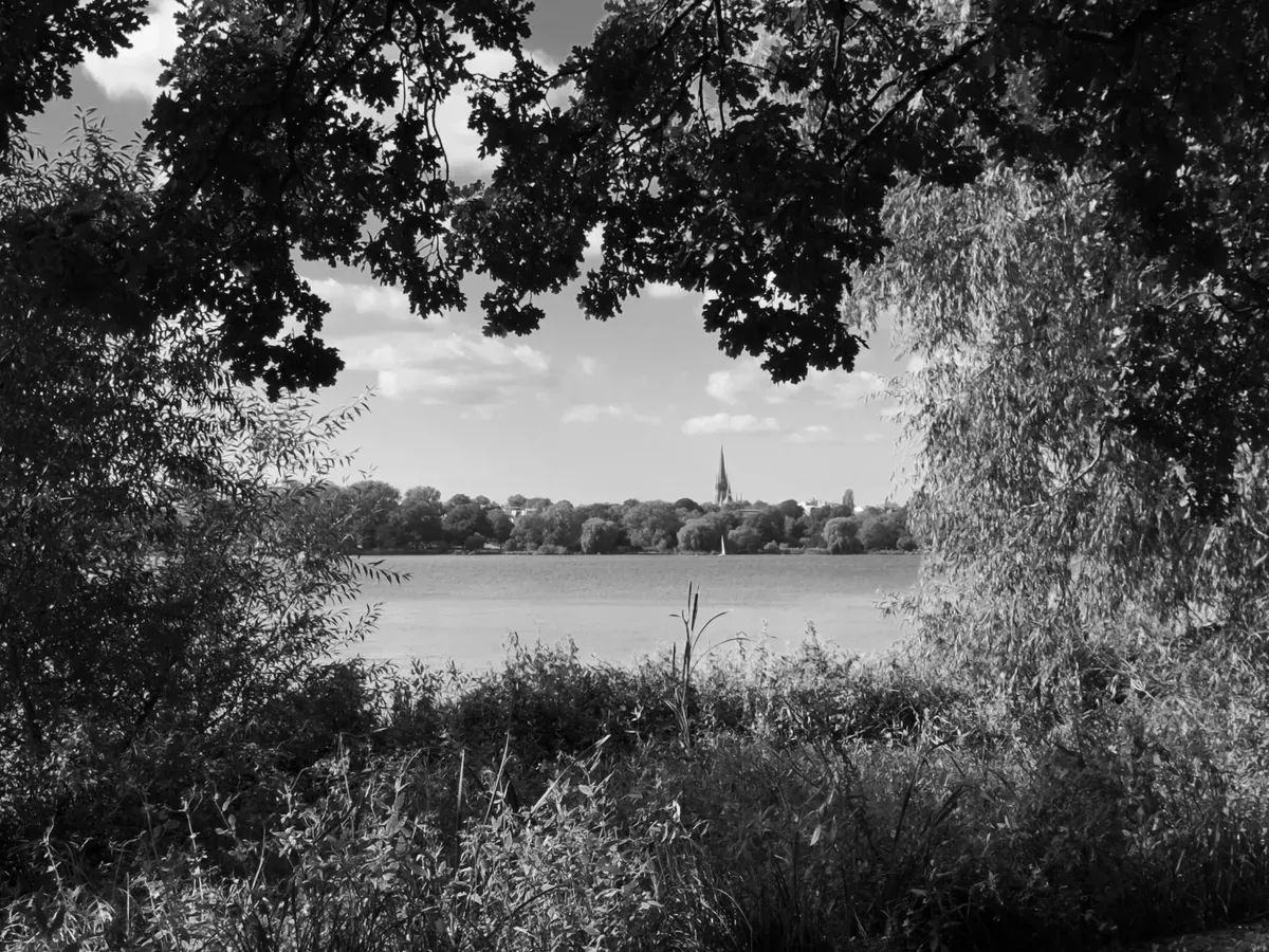 A serene black and white landscape featuring a lake framed by tree branches and foliage. In the distance, a steeple of a church is visible amidst a lush, wooded backdrop under a partly cloudy sky.