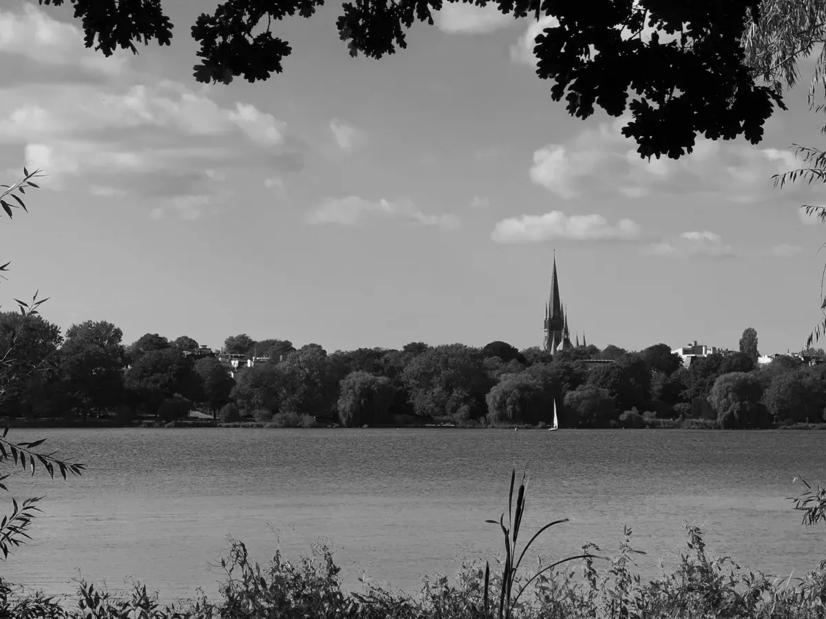 A serene lakeside scene in black and white, framed by foliage at the top. In the distance, a church spire rises above a line of trees along the shore, with a small sailboat visible on the water.