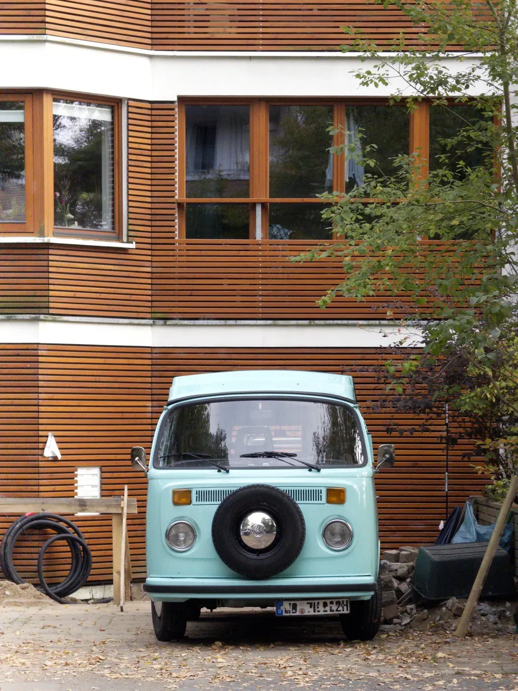 A light blue vintage van with a spare tire mounted on the front is parked in front of a modern wooden-paneled building. Surrounding the van are various items, including a green tarp and a stack of stones.